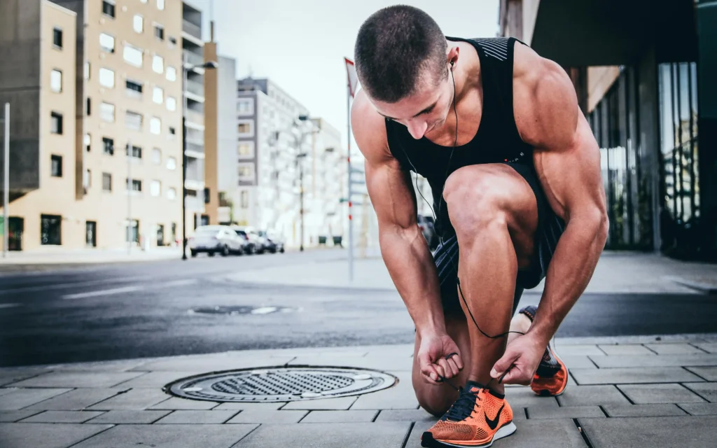 "A fit, muscular man in a black tank top tying his bright orange running shoes on a city sidewalk. He is wearing earphones and appears focused, preparing for a run in an urban environment with modern buildings in the background."