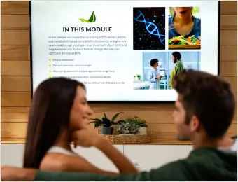 "A couple sits on a couch, viewing a television screen displaying a health and wellness module titled 'IN THIS MODULE.' The screen includes bullet points about nutrition, genetics, and lifestyle, along with related images. The couple appears engaged in the content, set in a cozy living room environment."