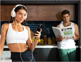 "A fit woman in a white sports bra and headphones smiles while holding her phone in a modern kitchen. In the background, a man in a tank top is reading The Ultimate Nutrition Bible, highlighting a focus on health, fitness, and nutrition."